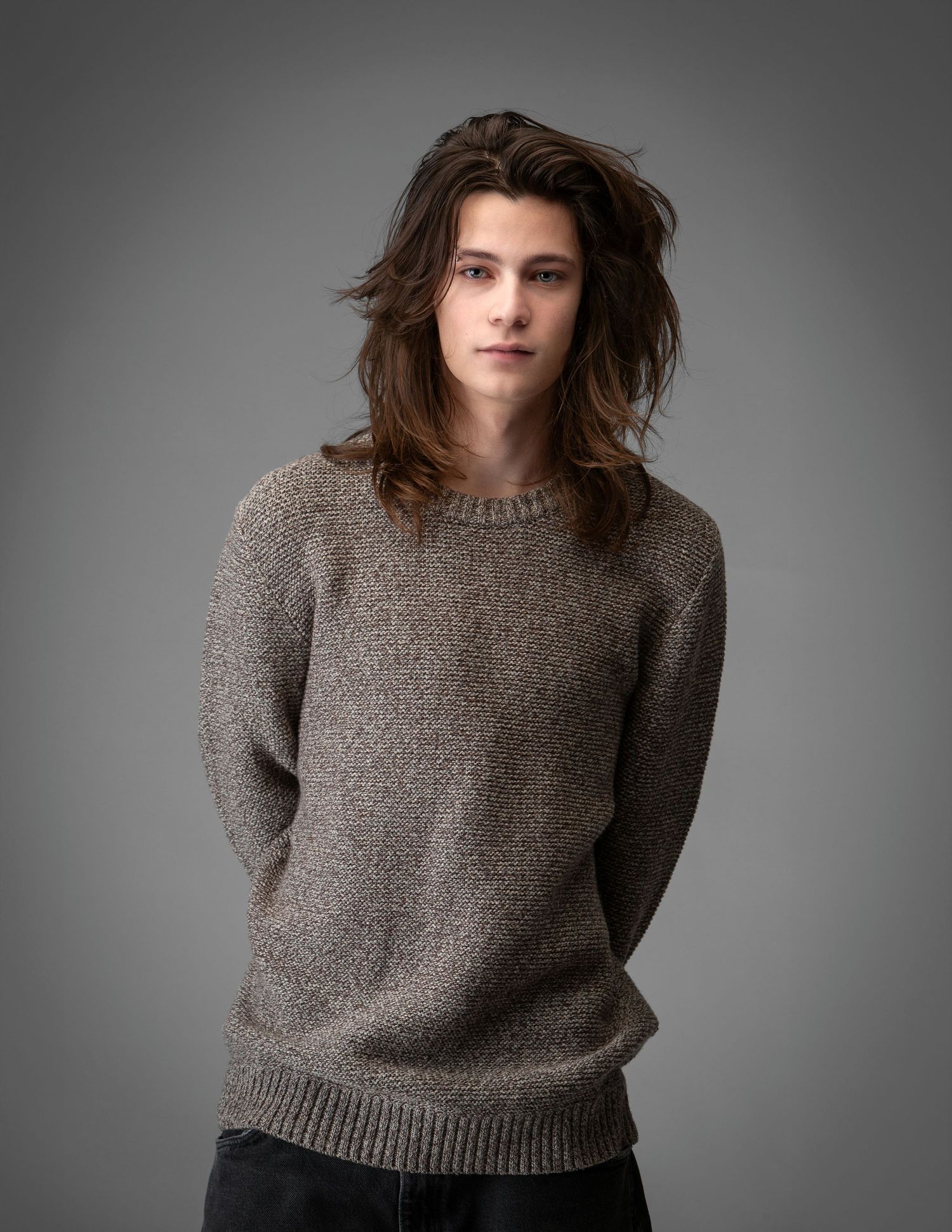 Fashion model wearing a gray knit sweater and dark pants poses against a neutral studio backdrop.