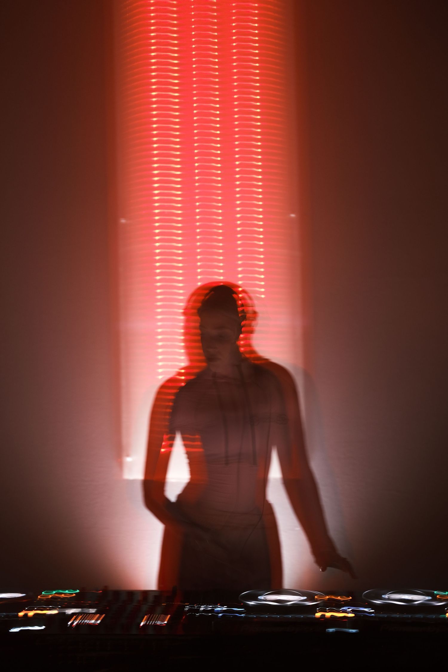 Silhouetted figure performing at DJ deck with dramatic red lighting and beams creating atmospheric effect.