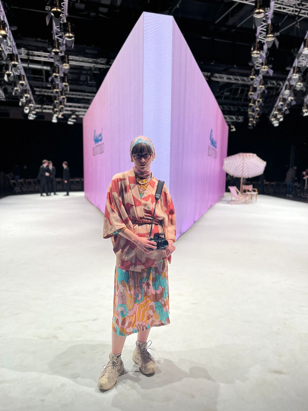 A person in a colorful patterned shirt and sunglasses poses on a white runway stage with pink lighting.