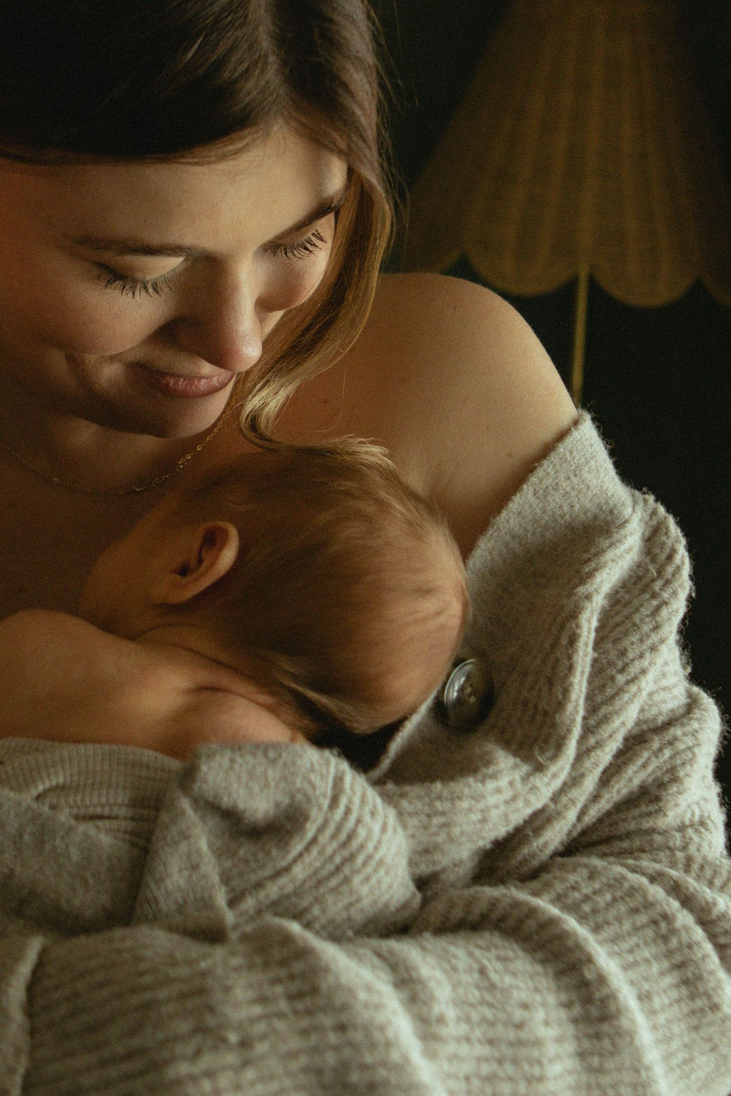 Smiling mother tenderly holding sleeping newborn baby wrapped in soft cream blanket.