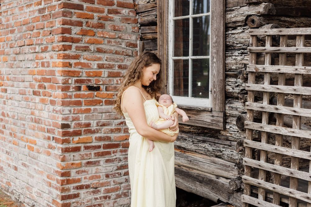 A mother wearing a pale yellow dress holds her baby girl wearing a matching dress in front of a brick fireplace outside a log cabin.