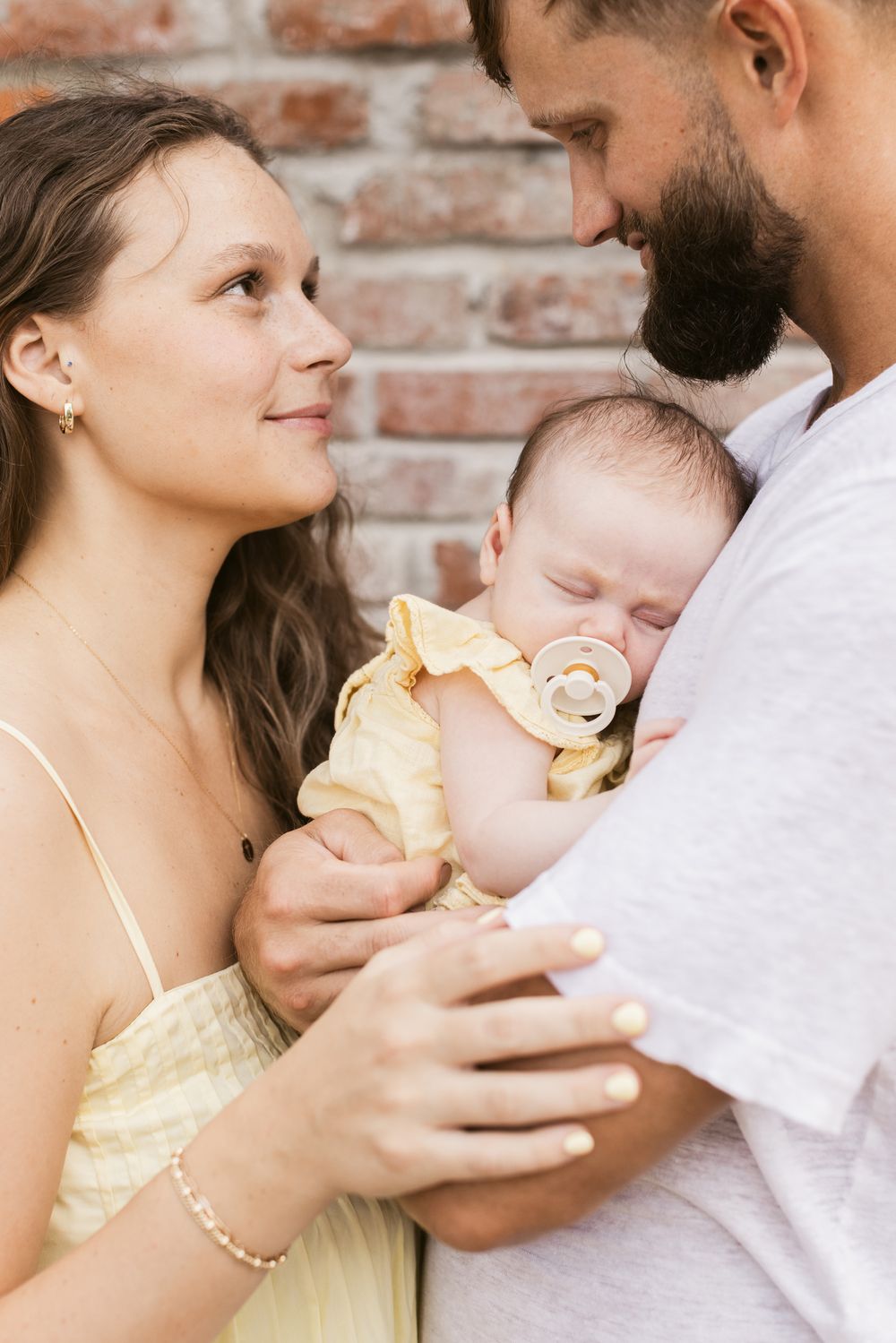 A mother wearing a pale yellow dress lovingly looks at the father holding their newborn baby girl in front of a brick fireplace.