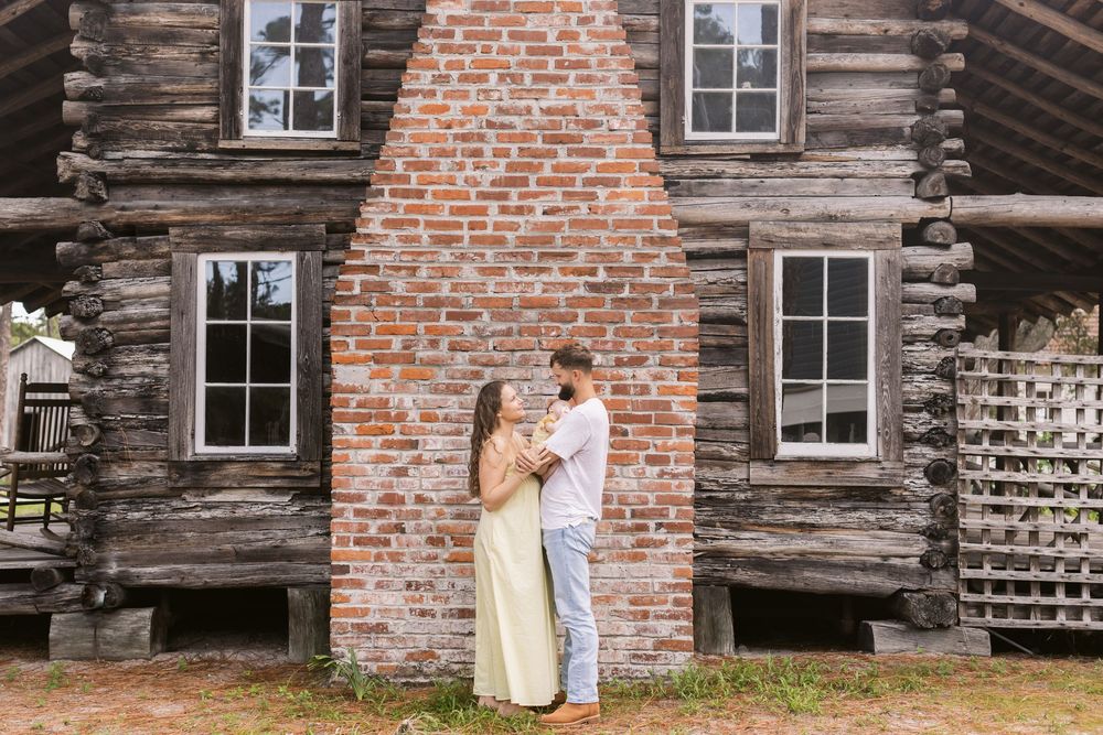 A coupe gazes at each other while holding their newborn daughter in front of a brick fireplace outside a log cabin for newborn photos.