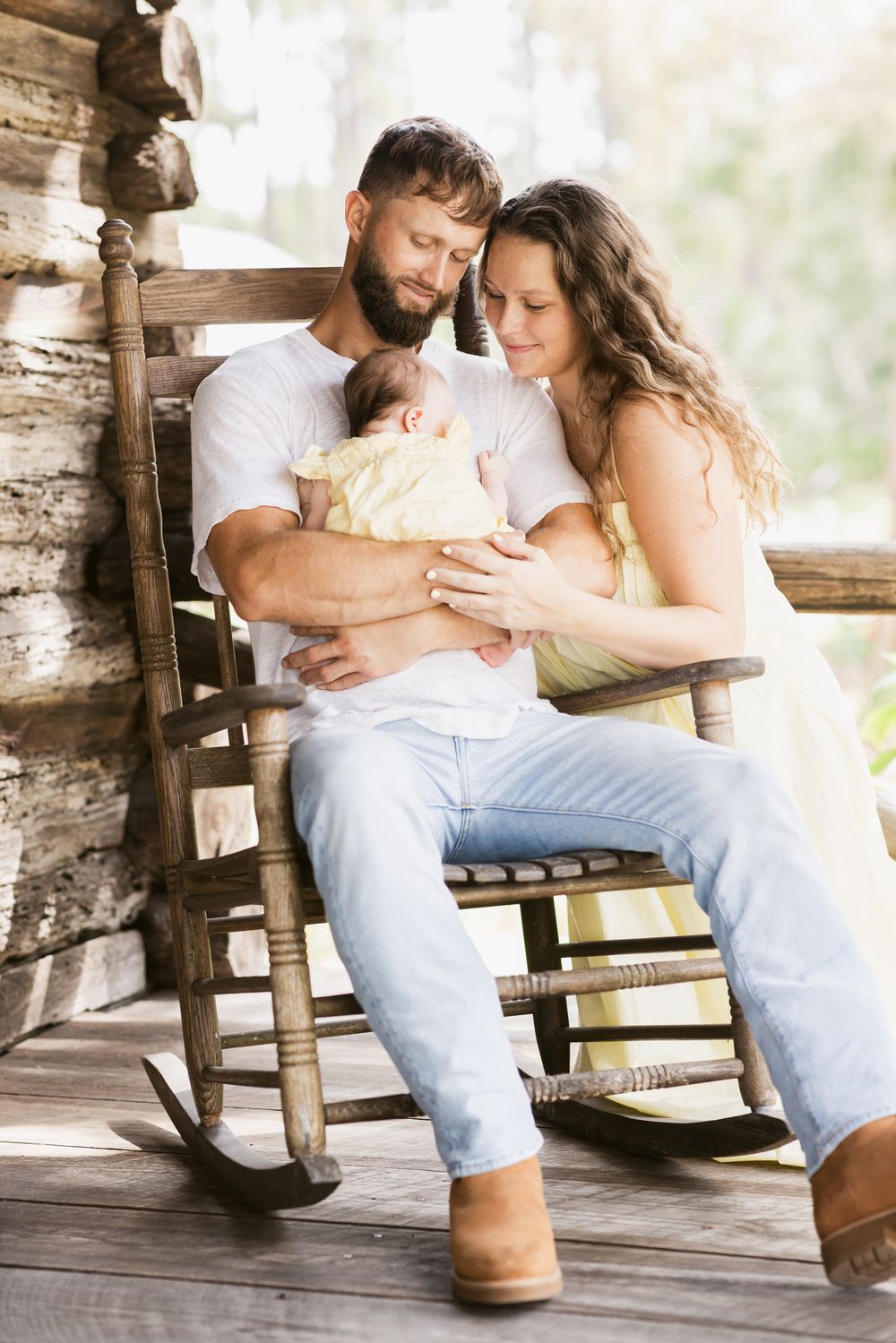 A father holds his newborn on his lap sitting on a rocking chair on a log cabin porch while the mother gazes at her newborn baby.