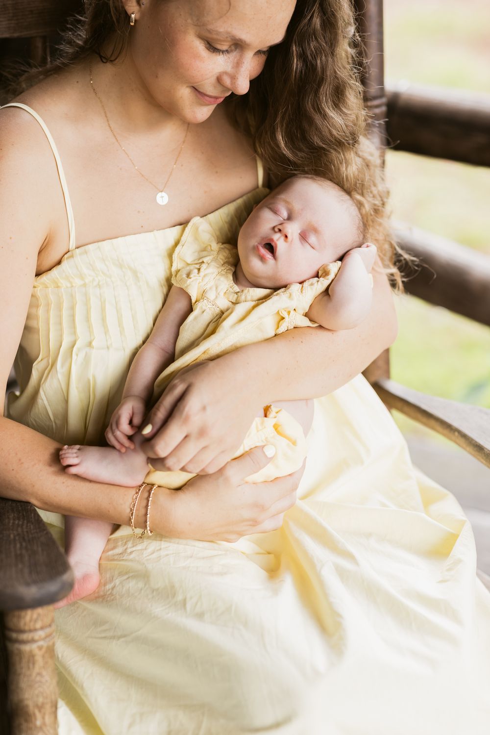 A mother in a pale yellow dress holds her newborn baby girl wearing a matching dress sitting on a rocking chair on a log cabin porch.