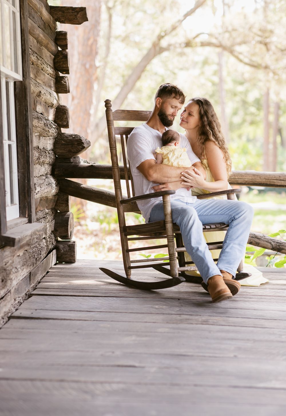 A mother gazes lovingly at the father who holds their newborn daughter on a rocking chair on a log cabin porch for newborn photos.