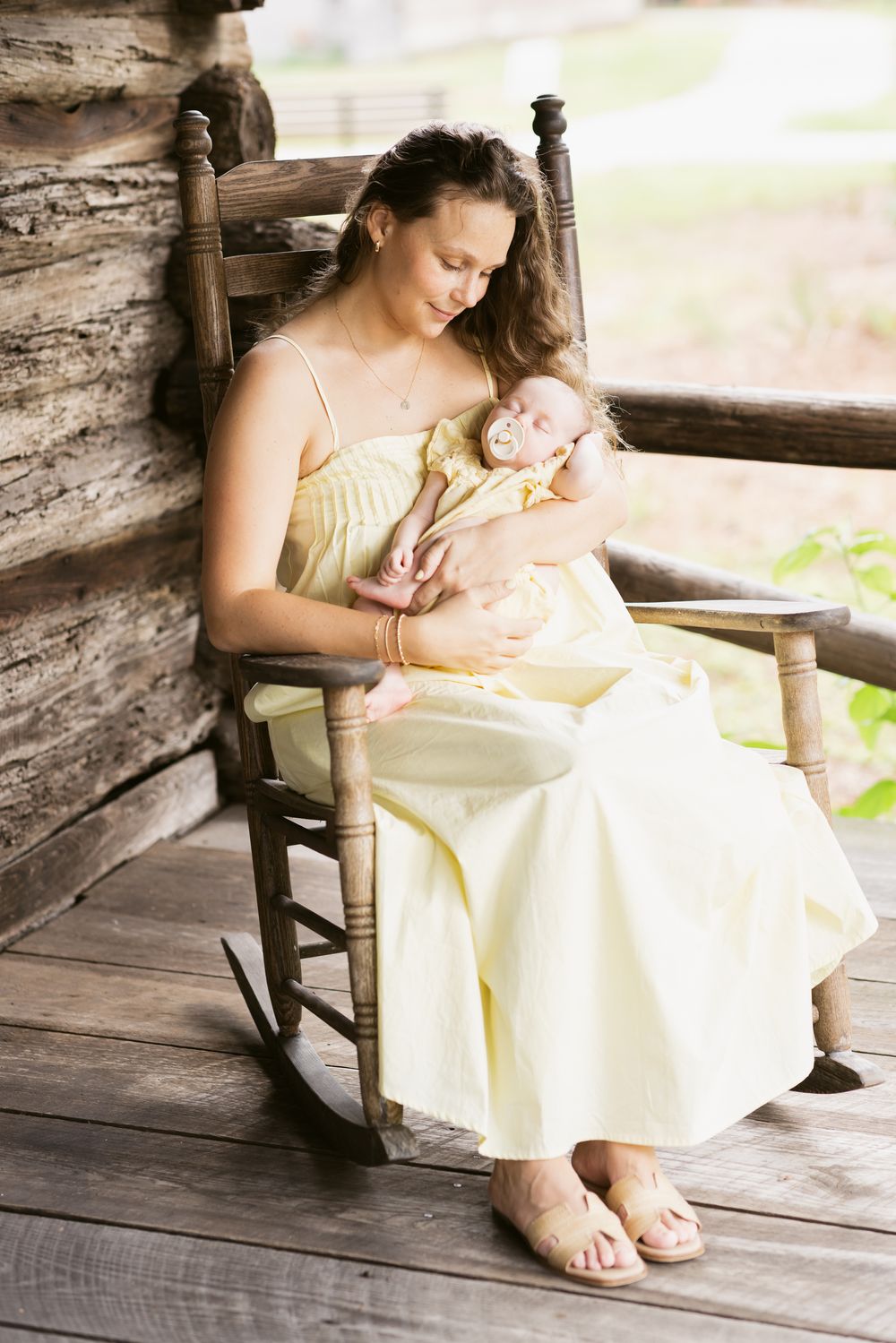 A mother and newborn baby girl wearing matching dresses sit on rocking chair on a log cabin porch. North Conway newborn photographer