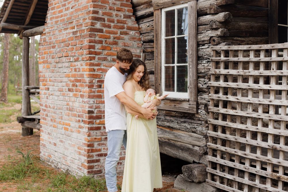 A father embraces the mother from behind holding their infant daughter in front of a brick fireplace outside a log cabin for newborn photos.