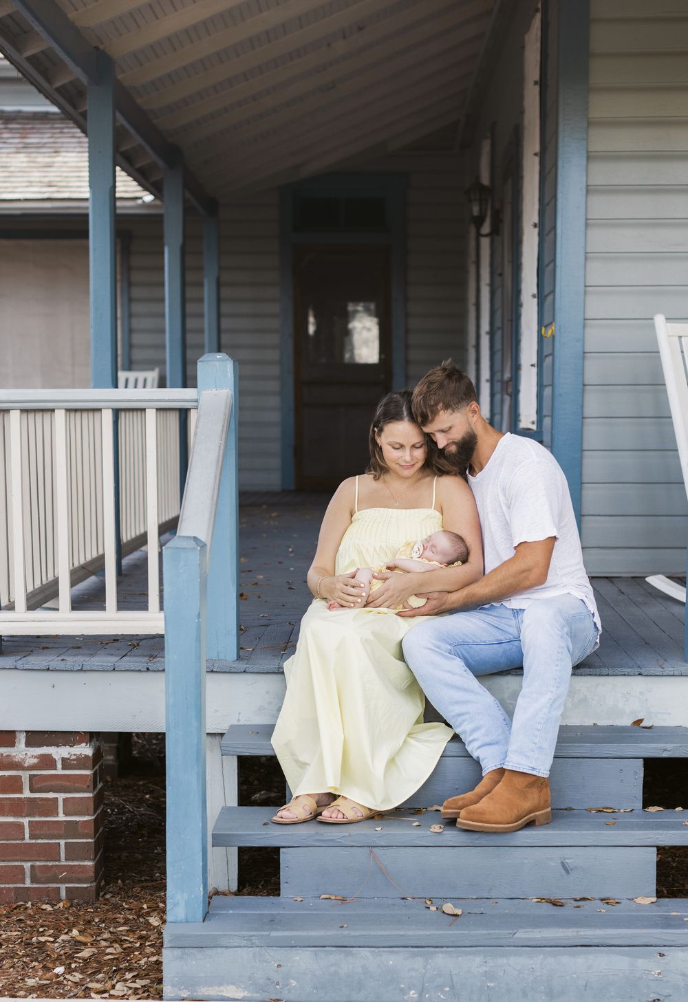 A couple holds their newborn gazing at her sitting on a pale blue farmer's porch for newborn photos. Dover, NH newborn photographer.
