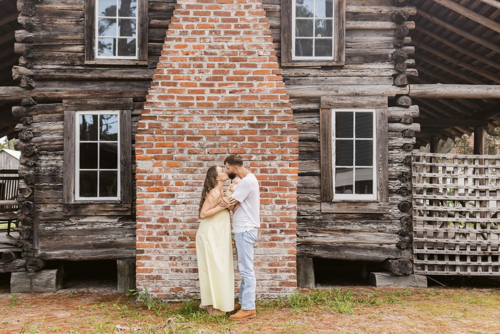 A couple kisses while holding their newborn in front of a brick fireplace outside a log cabin for a newborn photo shoot.