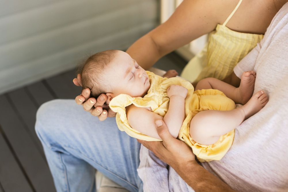 Newborn girl wearing yellow lays on her father's lap being also cradled by her mother on a farmer's porch. Alton Bay newborn photographer.