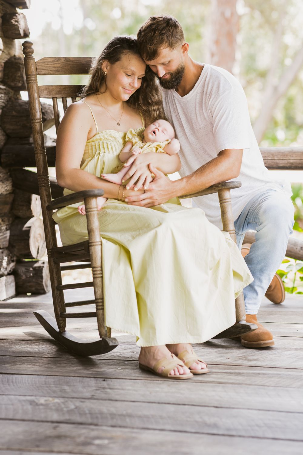 A mother siting on a rocking chair and father lovingly gaze at their newborn while on a log cabin porch for newborn photos.