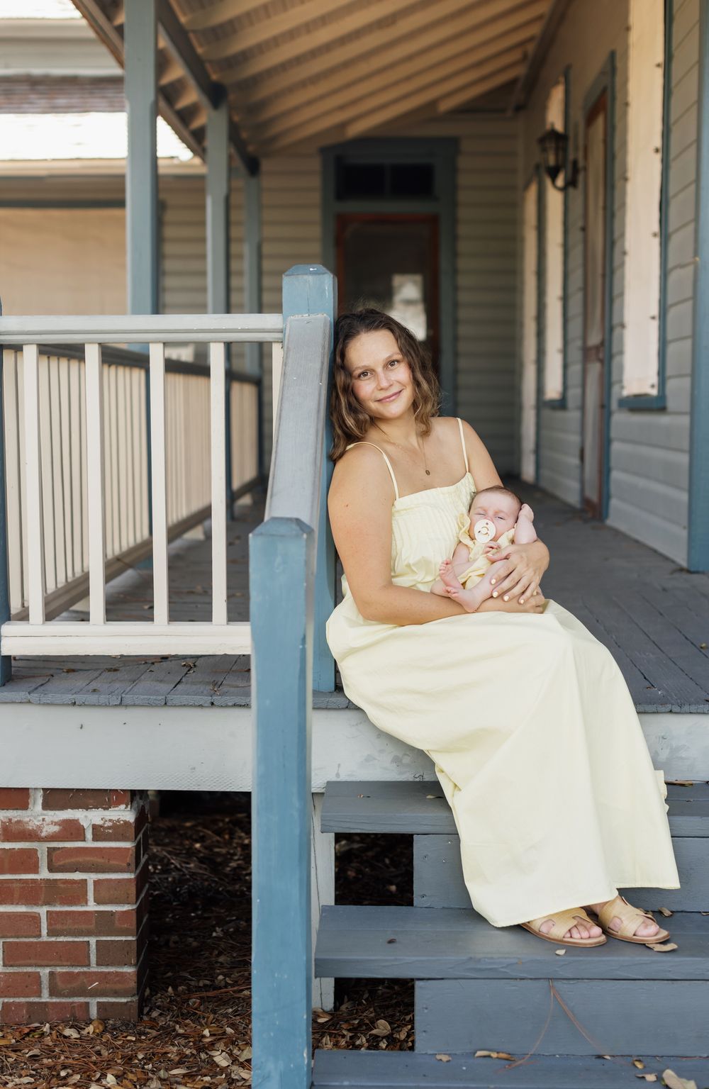 A mother wearing a pale yellow dress holds her newborn wearing a matching outfit siting on a pale blue porch for newborn photos.