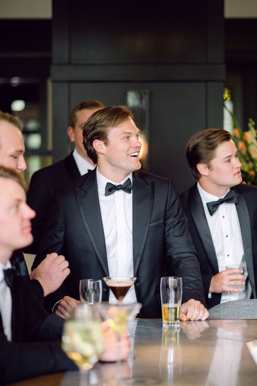 Groomsmen in tuxedos share laughs at a wedding reception while seated at a bar.
