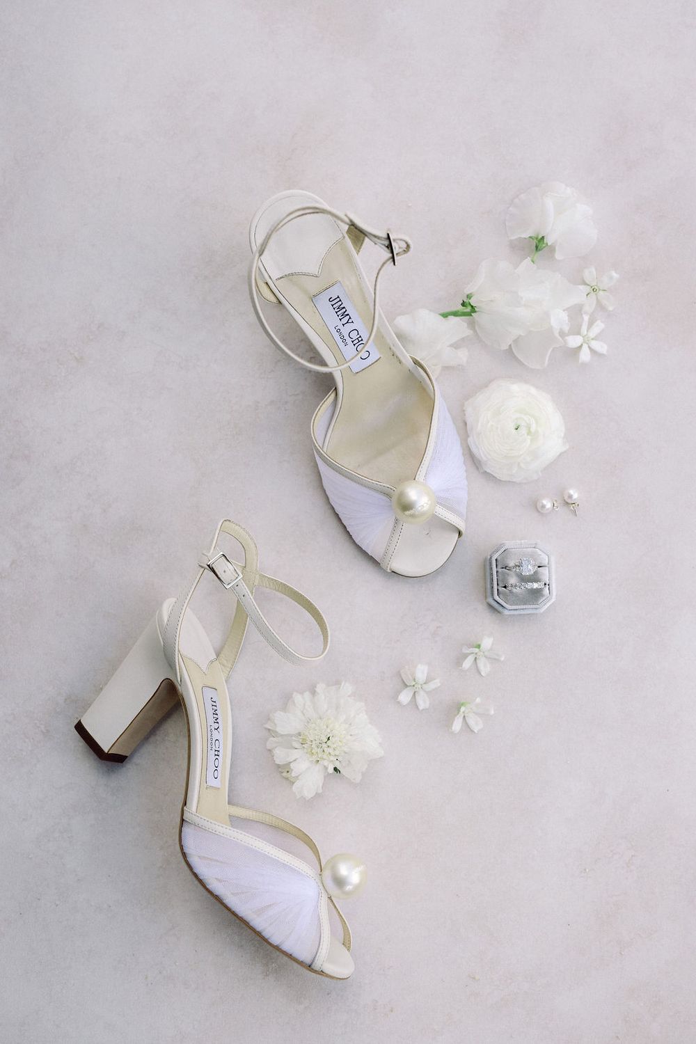 Elegant white bridal sandals with block heels surrounded by delicate white flowers and petals on a neutral background.