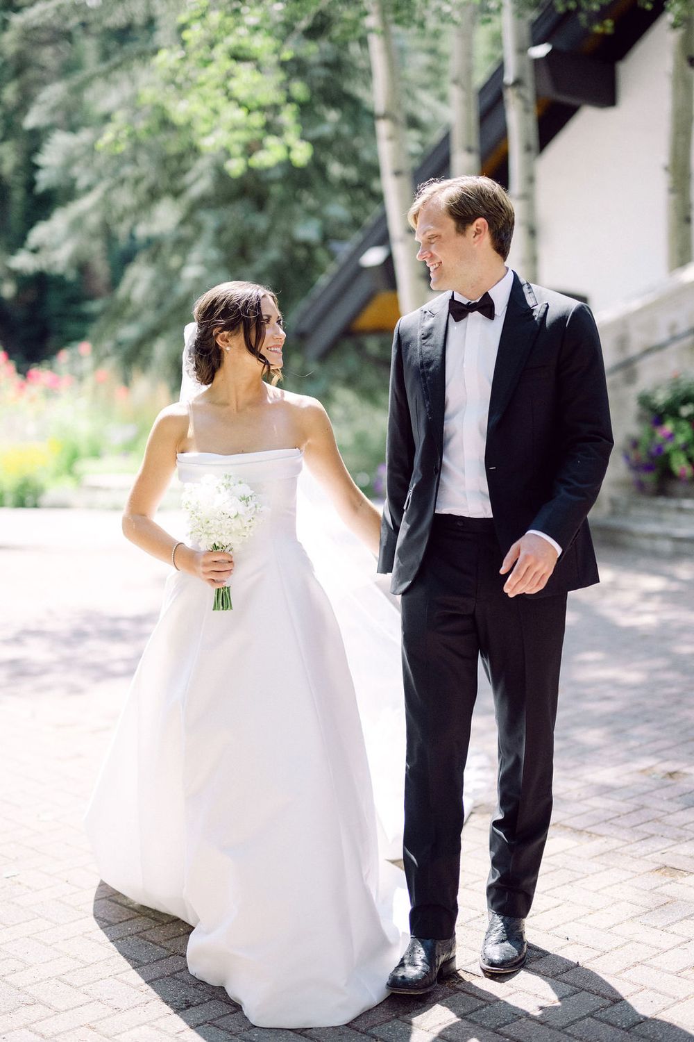 A bride in a white gown holding a bouquet stands with a groom in a black tuxedo on a stone path surrounded by greenery.