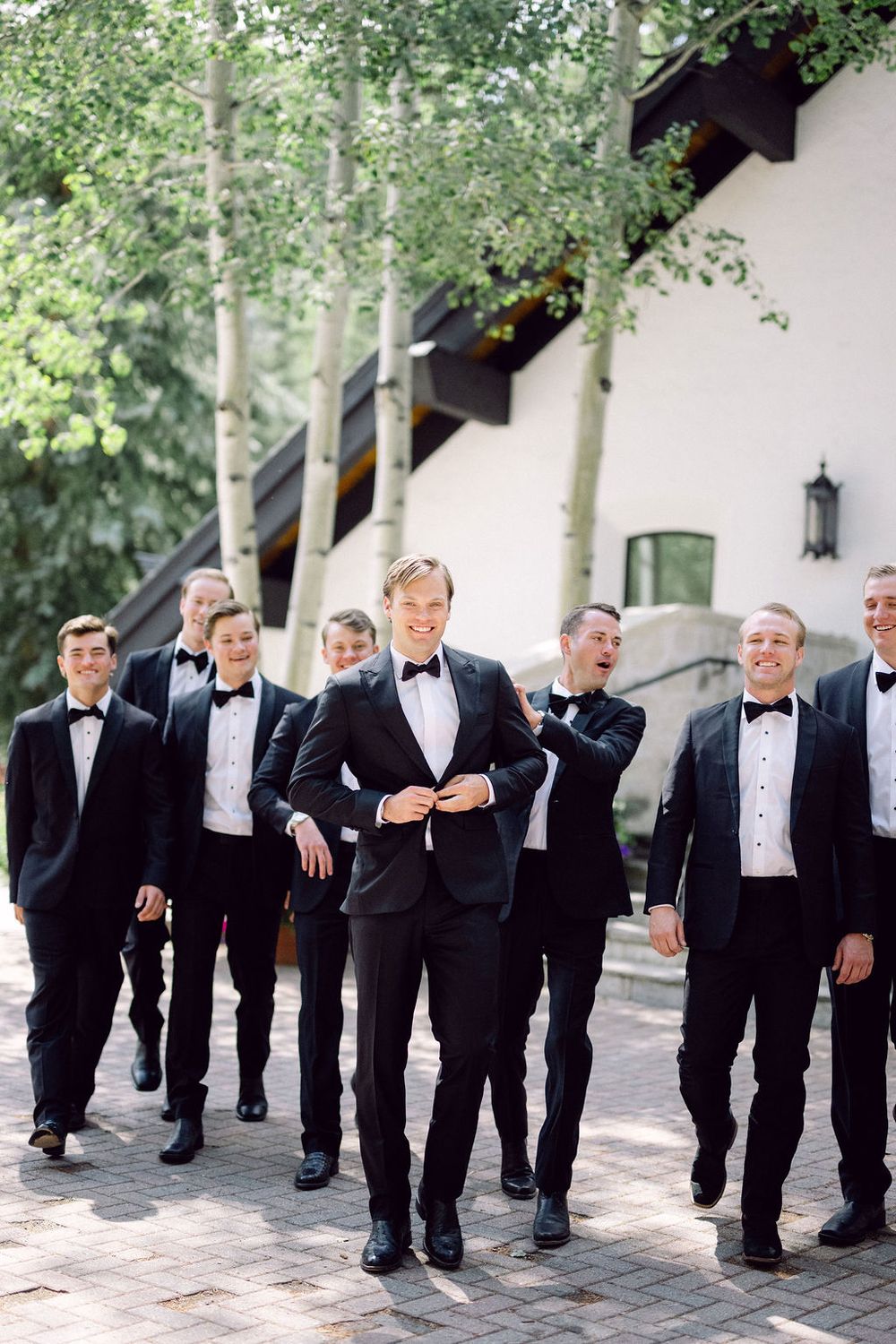 Groomsmen in black tuxedos pose casually against a white wall with trees in an artistic wedding photo series.