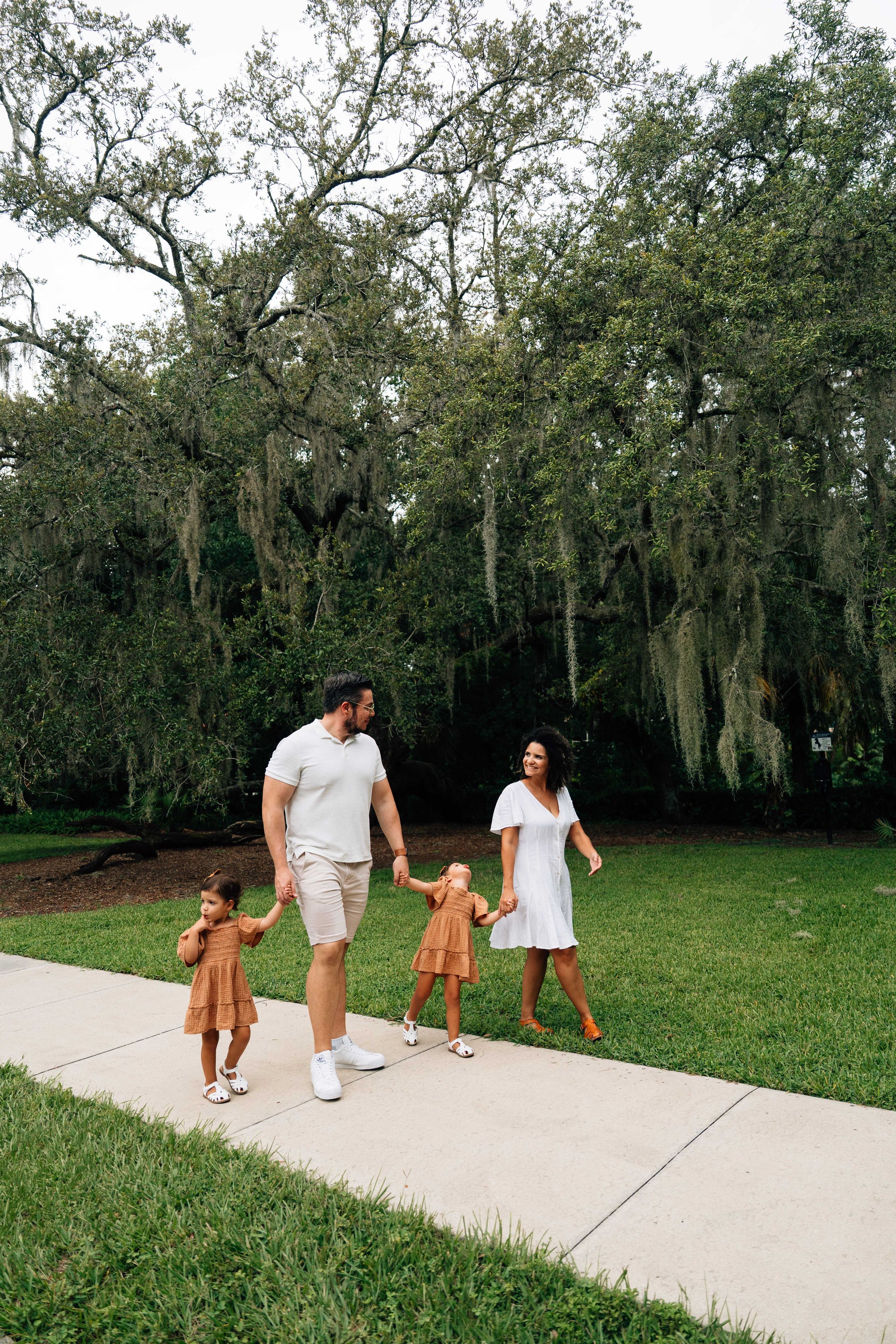 A family walks together on a concrete path through a grassy area with moss-covered trees in the background.
