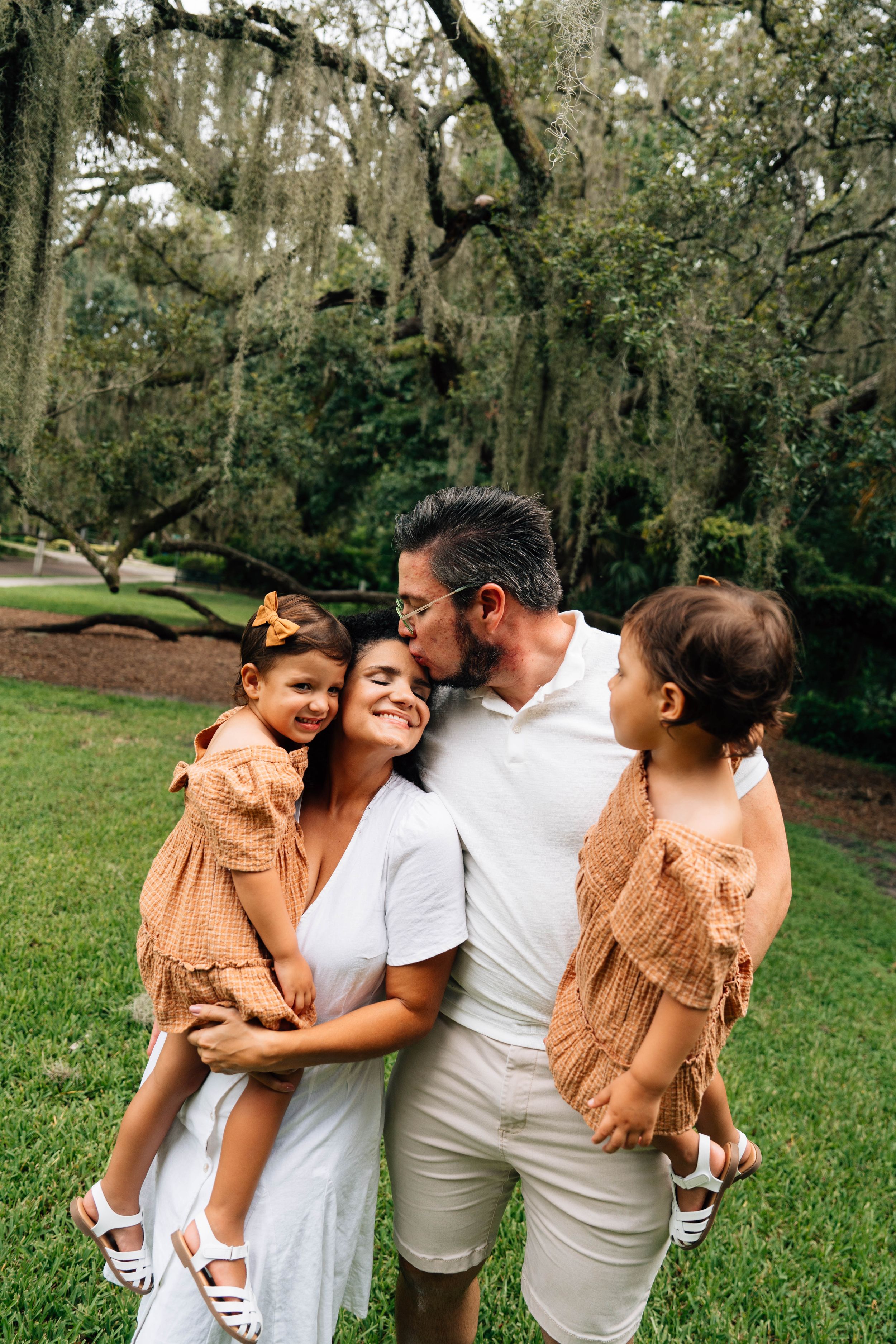 A family wearing coordinated white and beige outfits shares tender moments together in a lush outdoor setting.