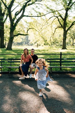 A young child runs joyfully on a sunlit path in Central Park while two people watch from a bench in the background.
