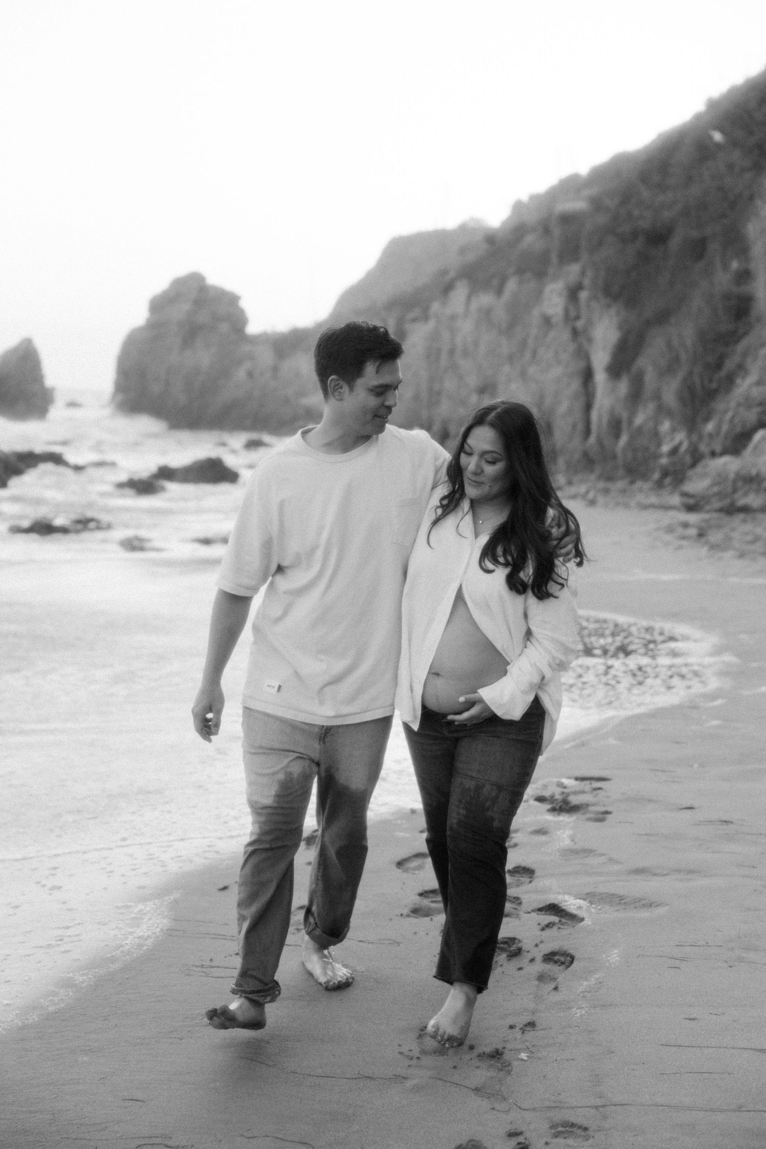 A couple walks barefoot along a rocky beach with cliffs in the background, captured in black and white.