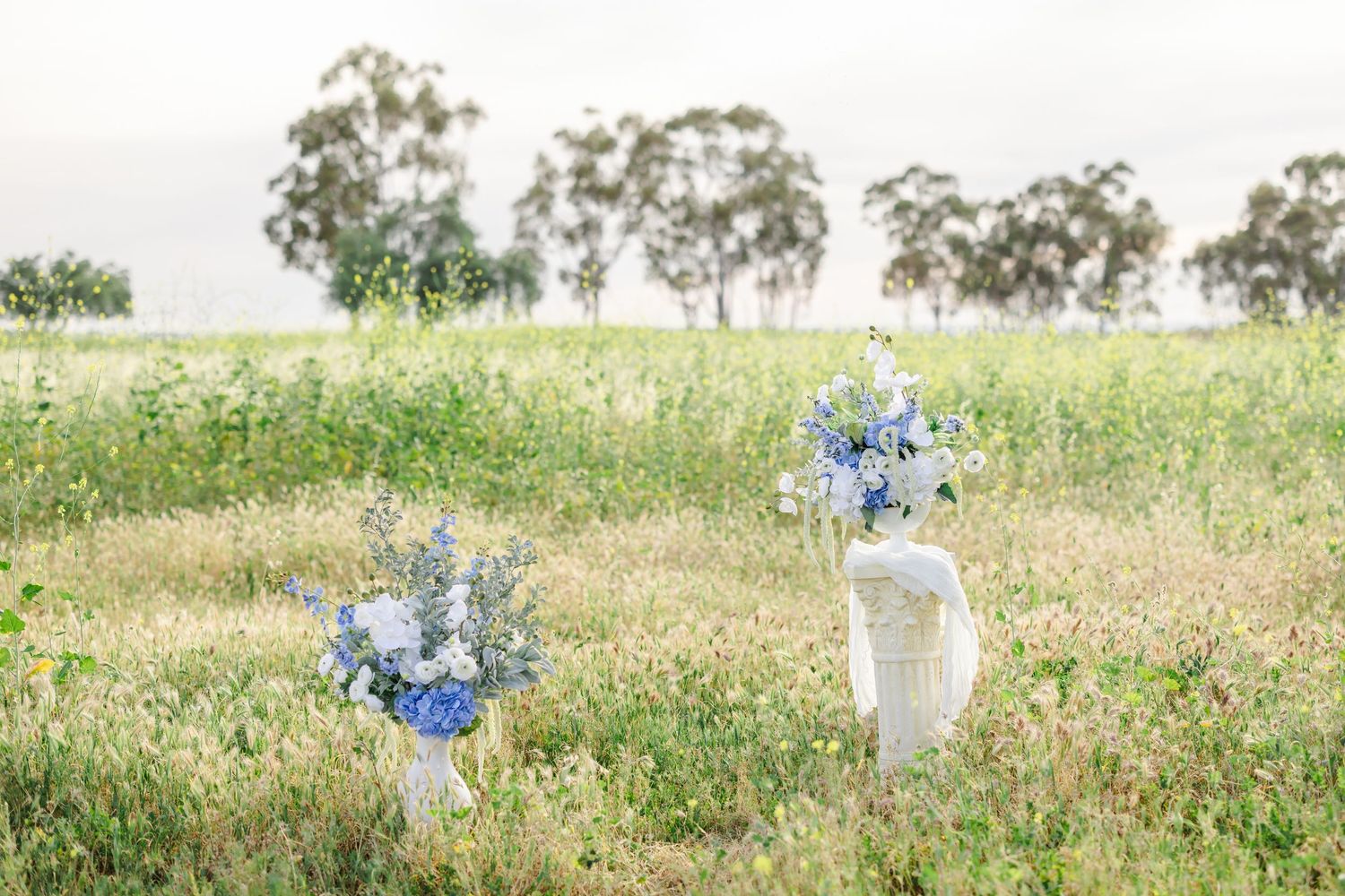 Scenic outdoor setting with white pillars and blue floral arrangements in a natural meadow.