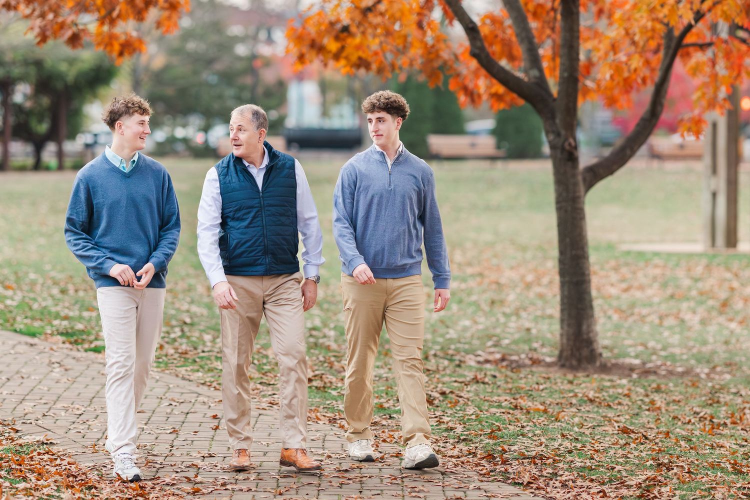 Three people walk together on fallen autumn leaves beneath vibrant orange maple trees in a park setting.