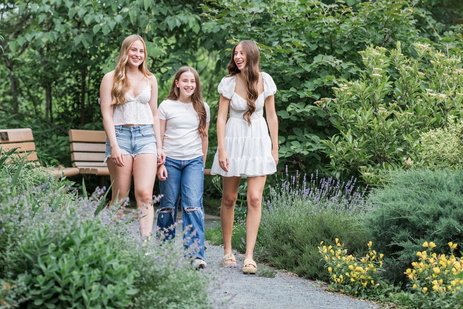 Three people walk together on a garden path surrounded by lush greenery and blooming flowers on a sunny day.