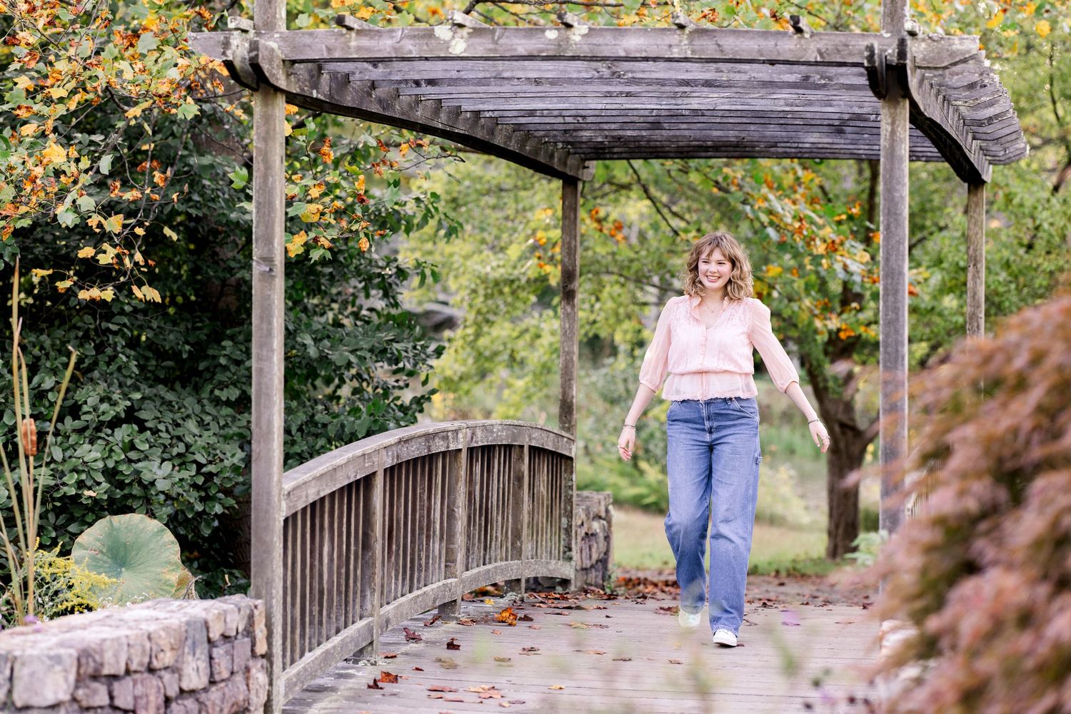 A person walks across a rustic covered wooden footbridge in a garden setting on an autumn day.
