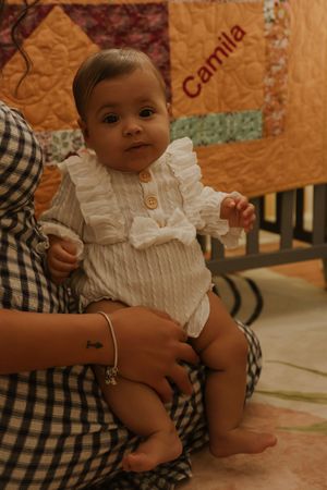 Baby in white ruffled outfit sits with parent in front of colorful quilted background.