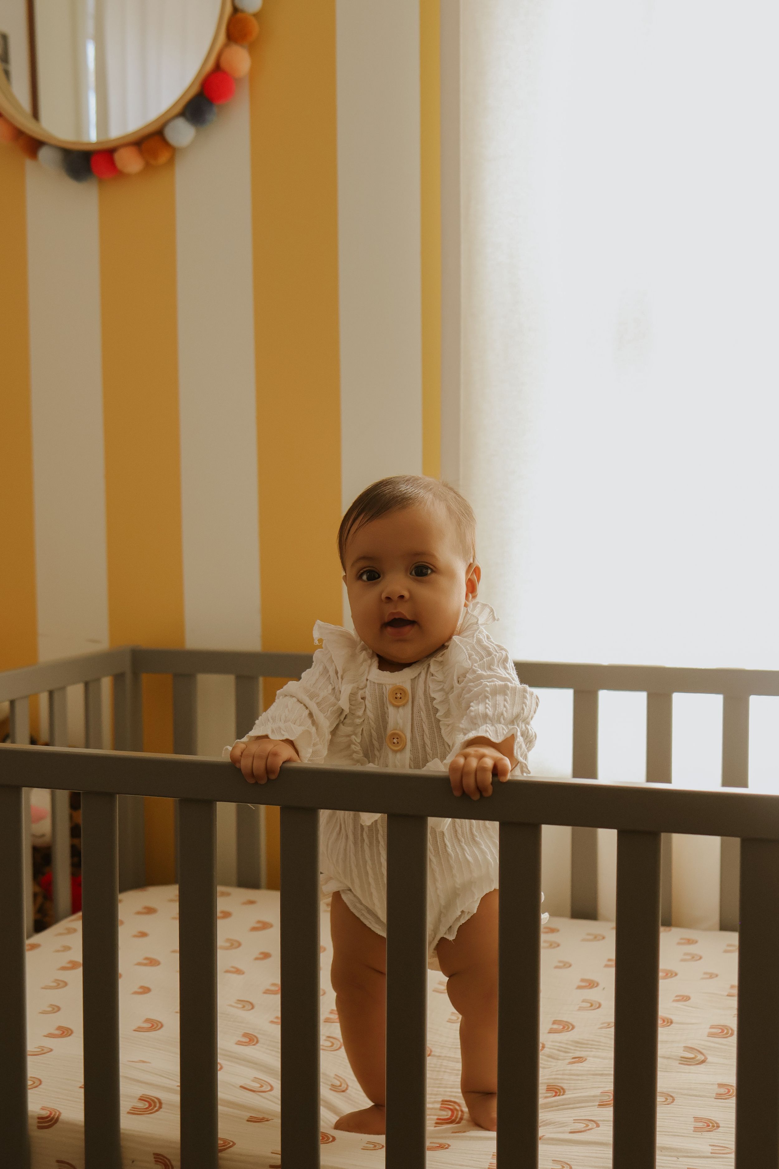 Happy baby in white outfit standing in gray crib with patterned sheets.