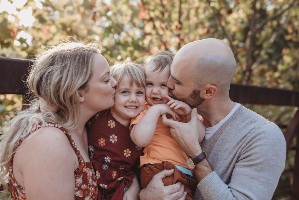Two parents kissing their smiling toddlers on the cheeks during a warm autumn family portrait.
