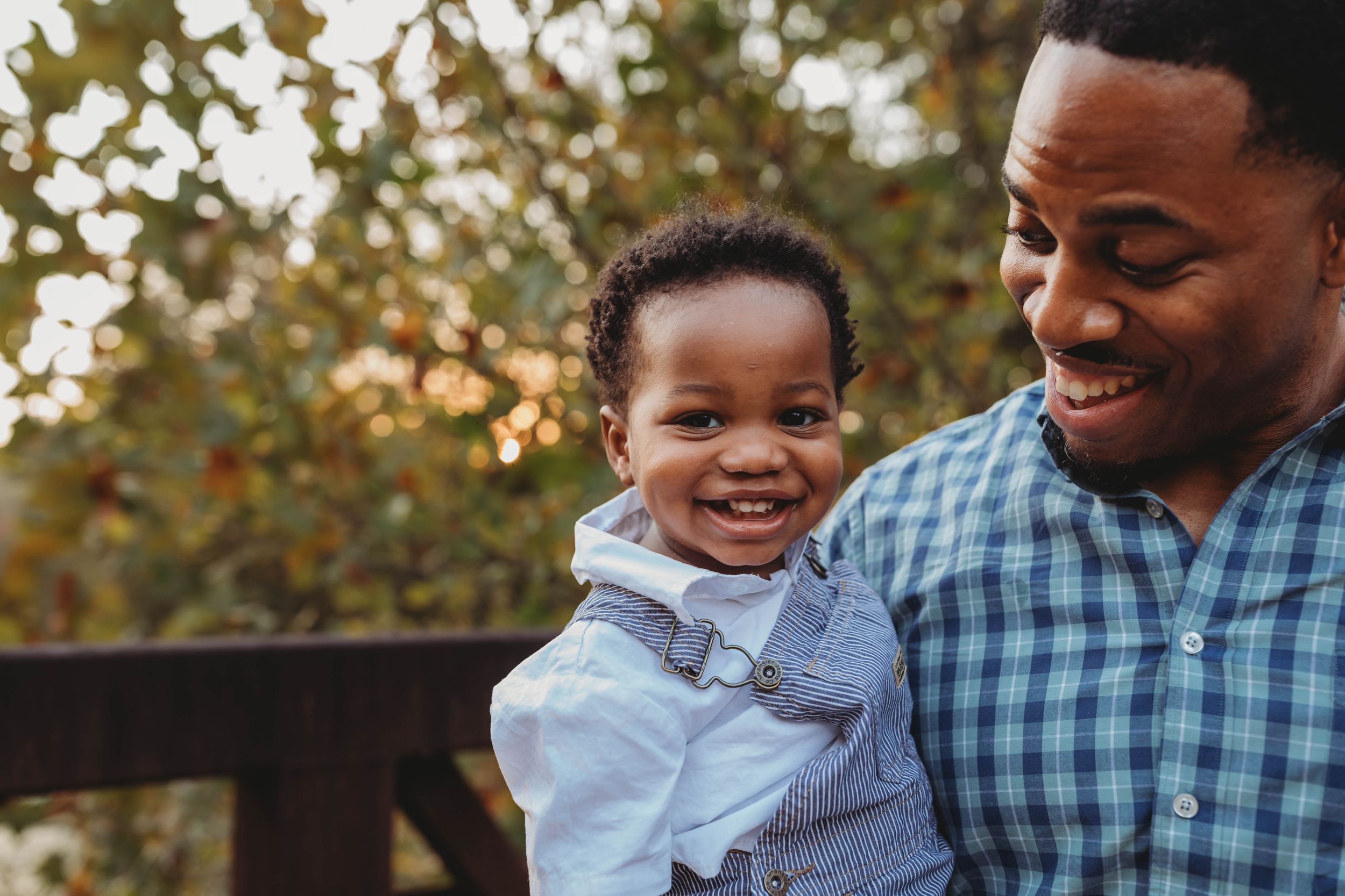 A smiling father holding his happy baby boy outdoors with autumn foliage in the background.