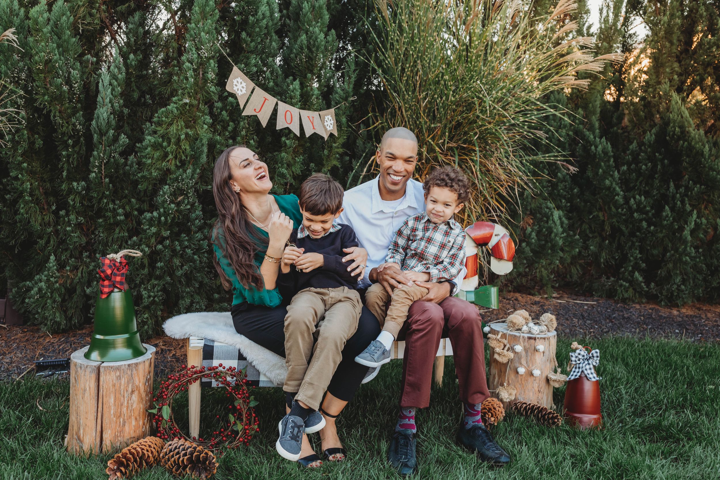 Happy family of four laughing together outdoors during a festive holiday photo session with pine trees in background.