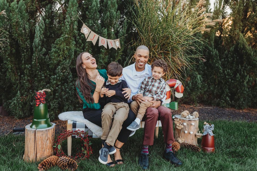 Happy family of four laughing together outdoors during a festive holiday photo session with pine trees in background.