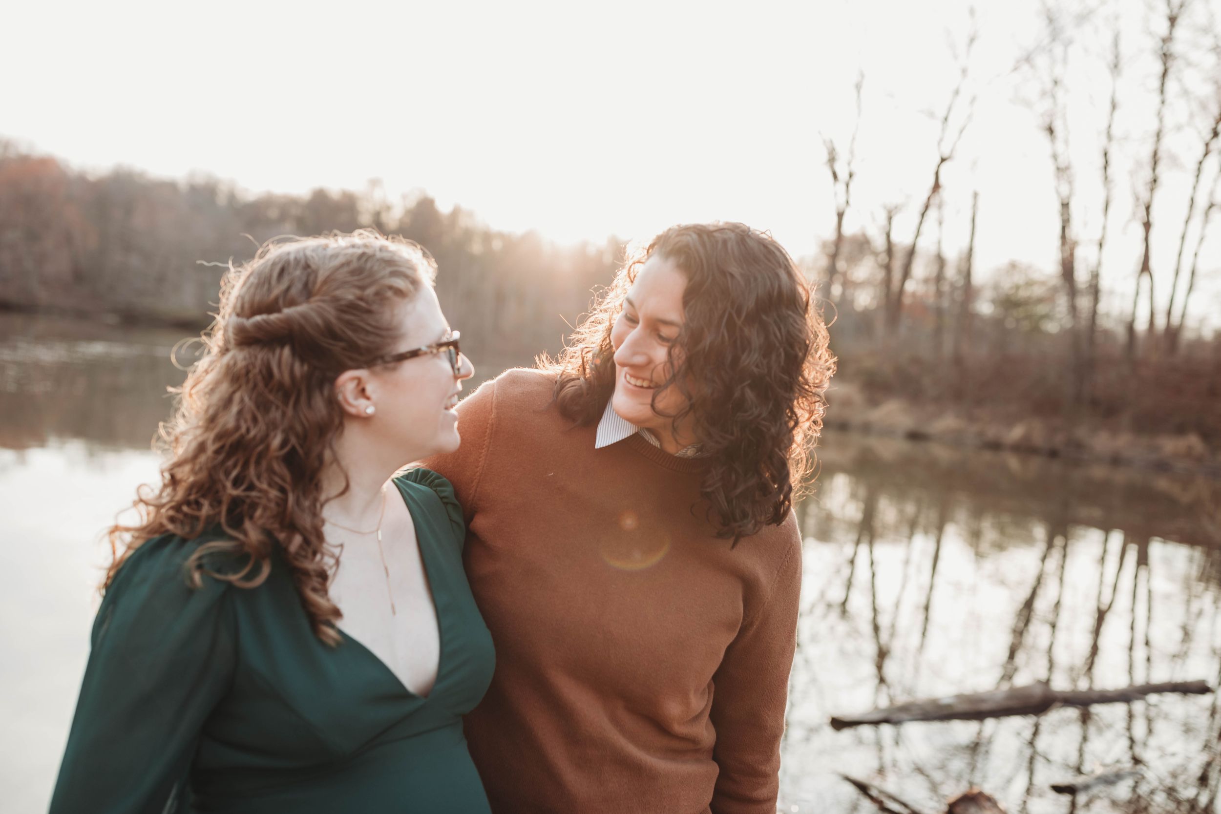 Two women smiling at each other near a sunlit lake during an outdoor portrait session.