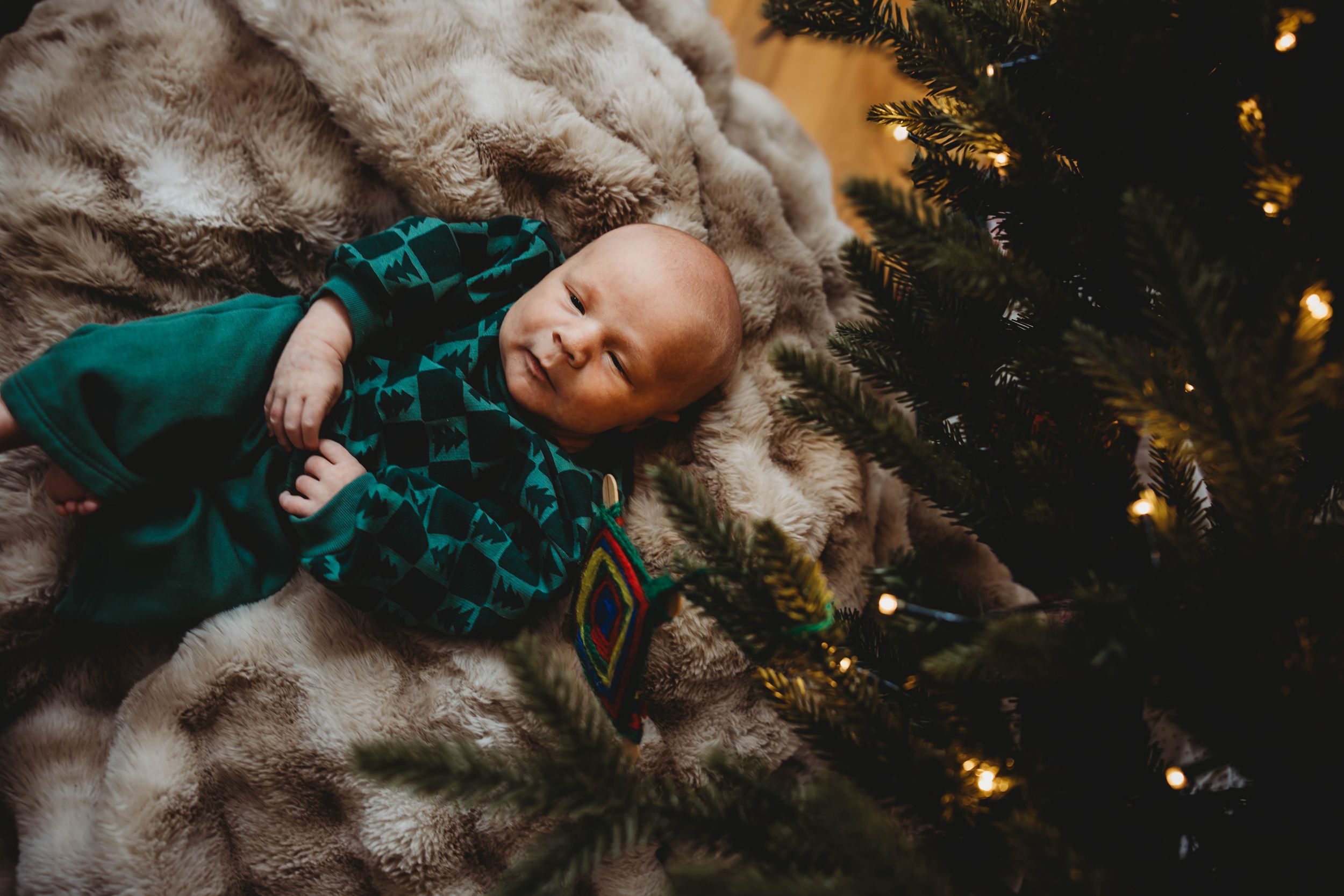 A smiling newborn baby in a green outfit lying on a fur blanket beside a Christmas tree.