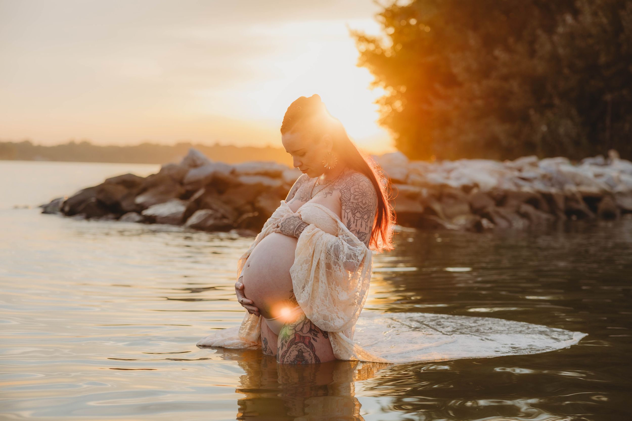 A pregnant woman in a white lace dress standing in water during a golden sunset maternity shoot.