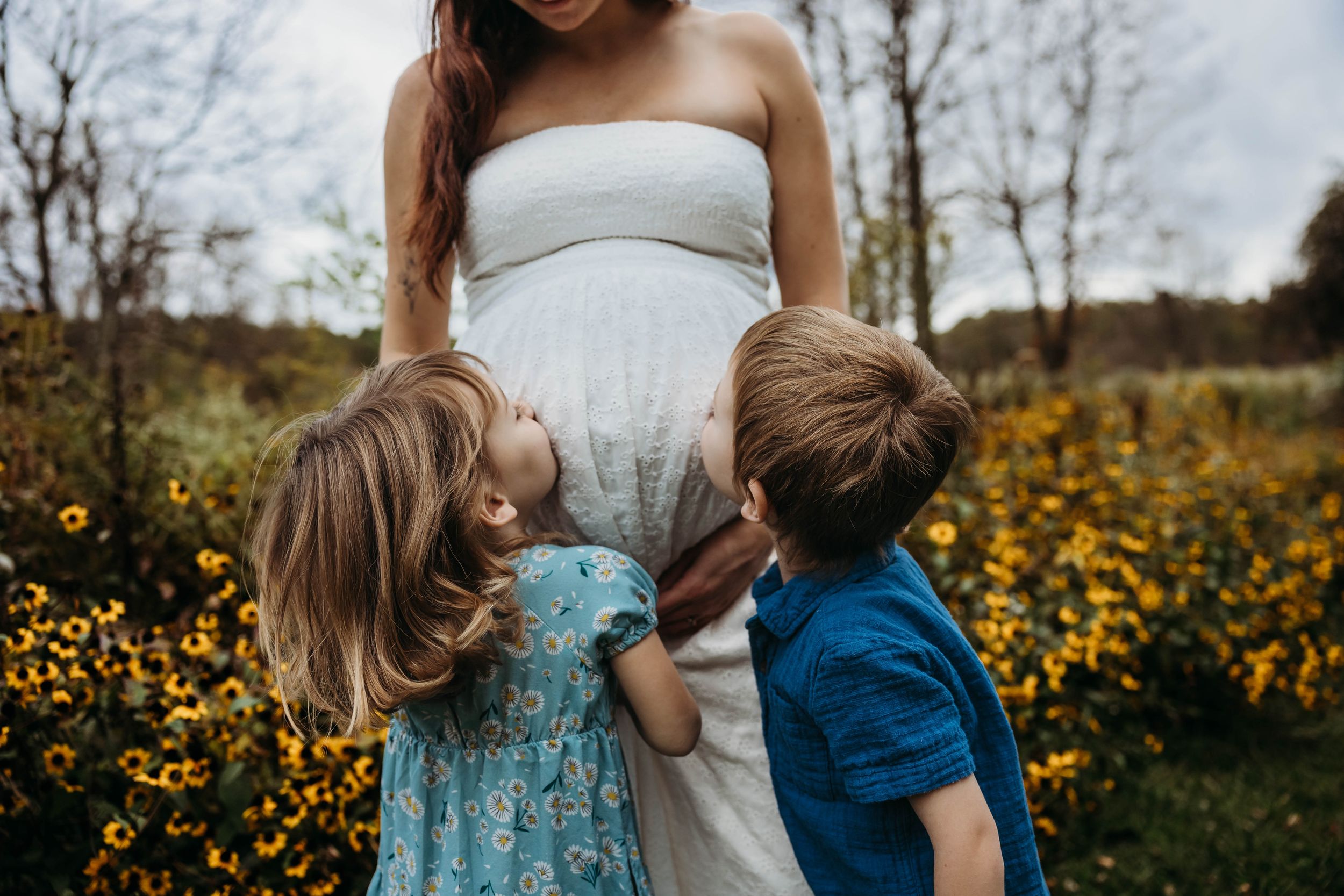 Two young children hugging their pregnant mothers belly in a field of yellow wildflowers.