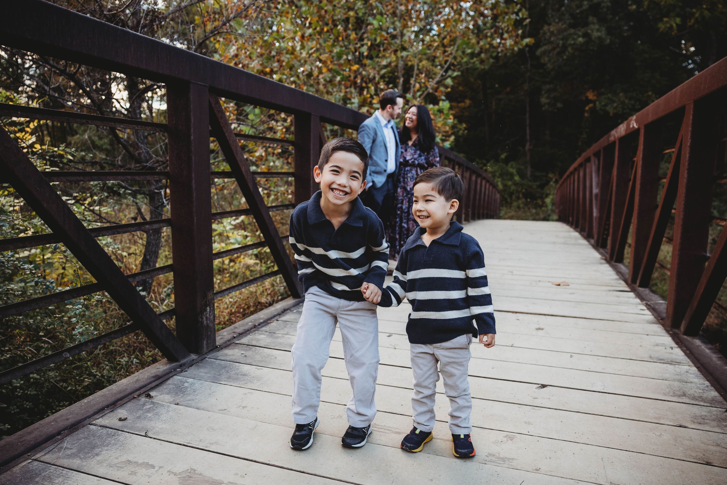 Two young brothers in matching striped sweaters holding hands and smiling on a wooden bridge during fall family photos.