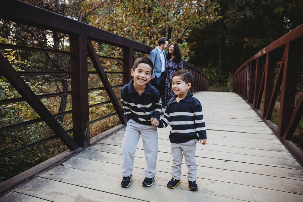 Two young brothers in matching striped sweaters holding hands and smiling on a wooden bridge during fall family photos.