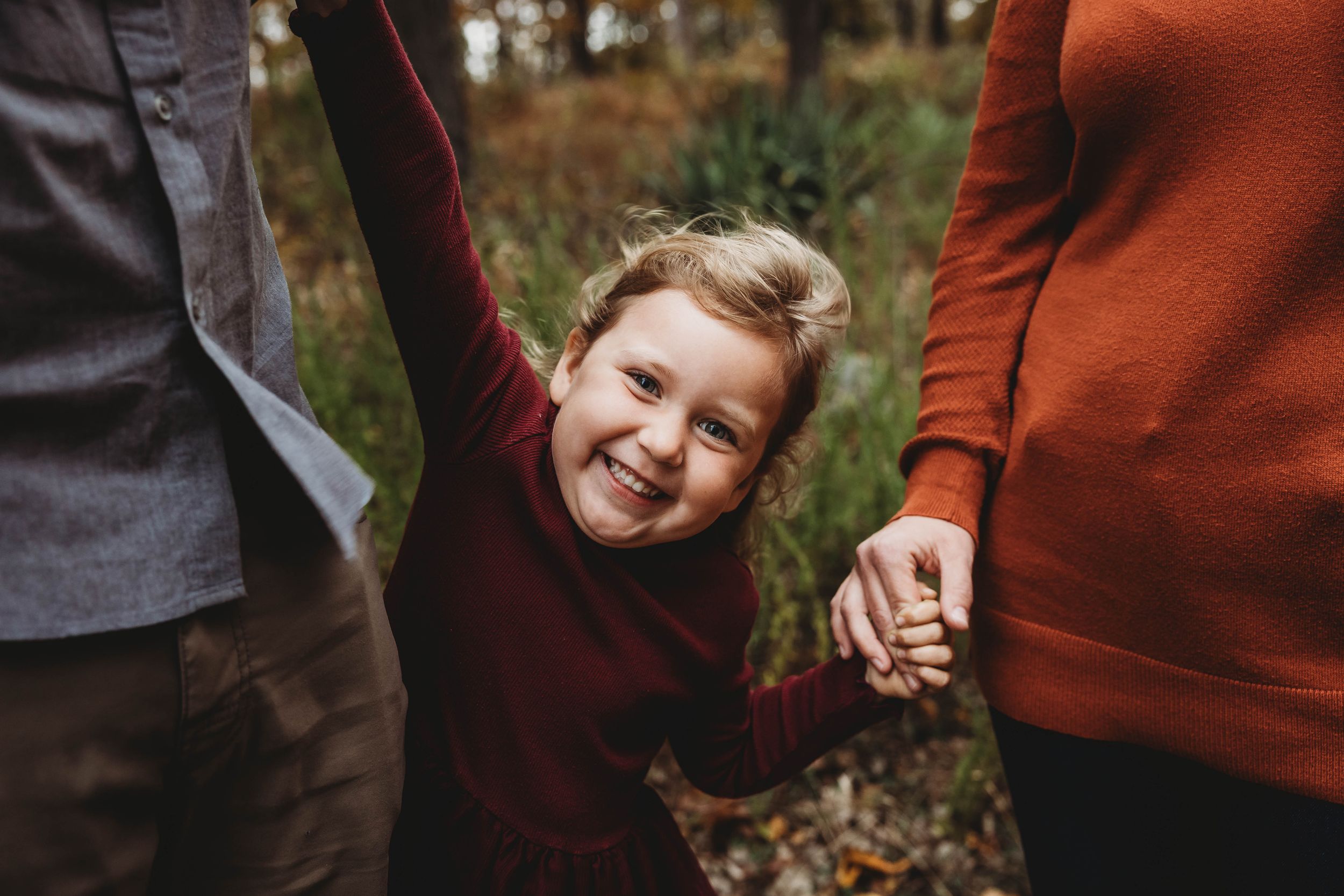 A joyful child swinging between two parents while holding their hands in an autumn forest.