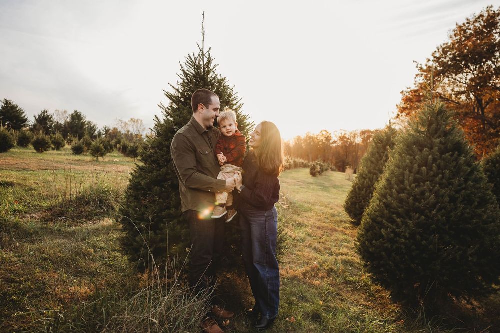 A family of three embracing warmly among Christmas trees at a tree farm during golden hour.