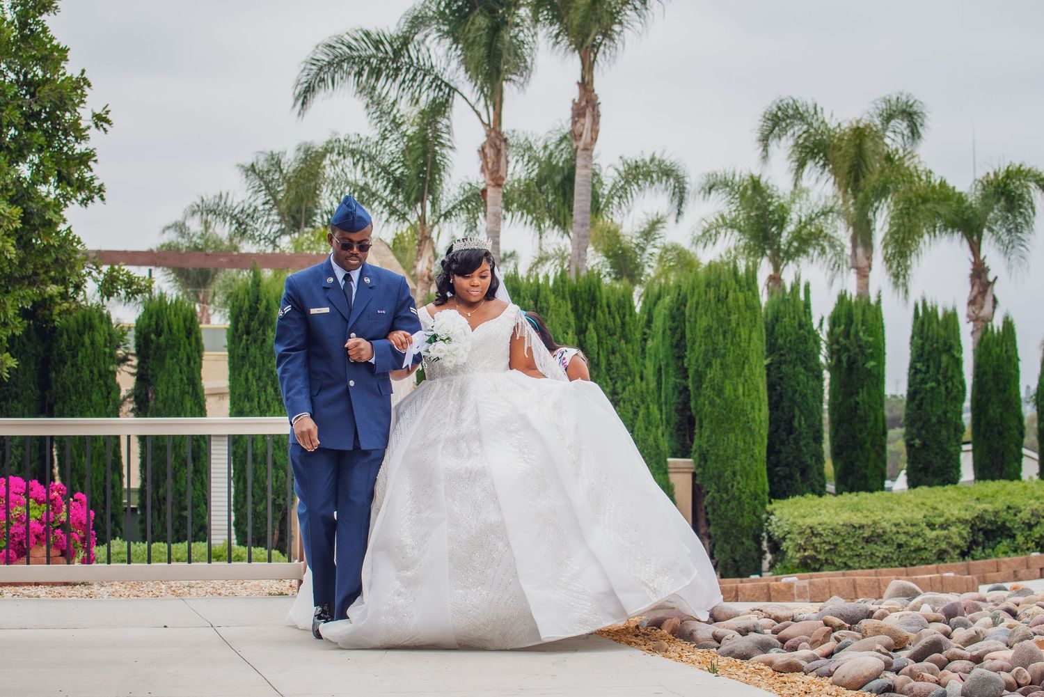 A couple in formal wedding attire walks together on a stone pathway surrounded by palm trees and greenery.