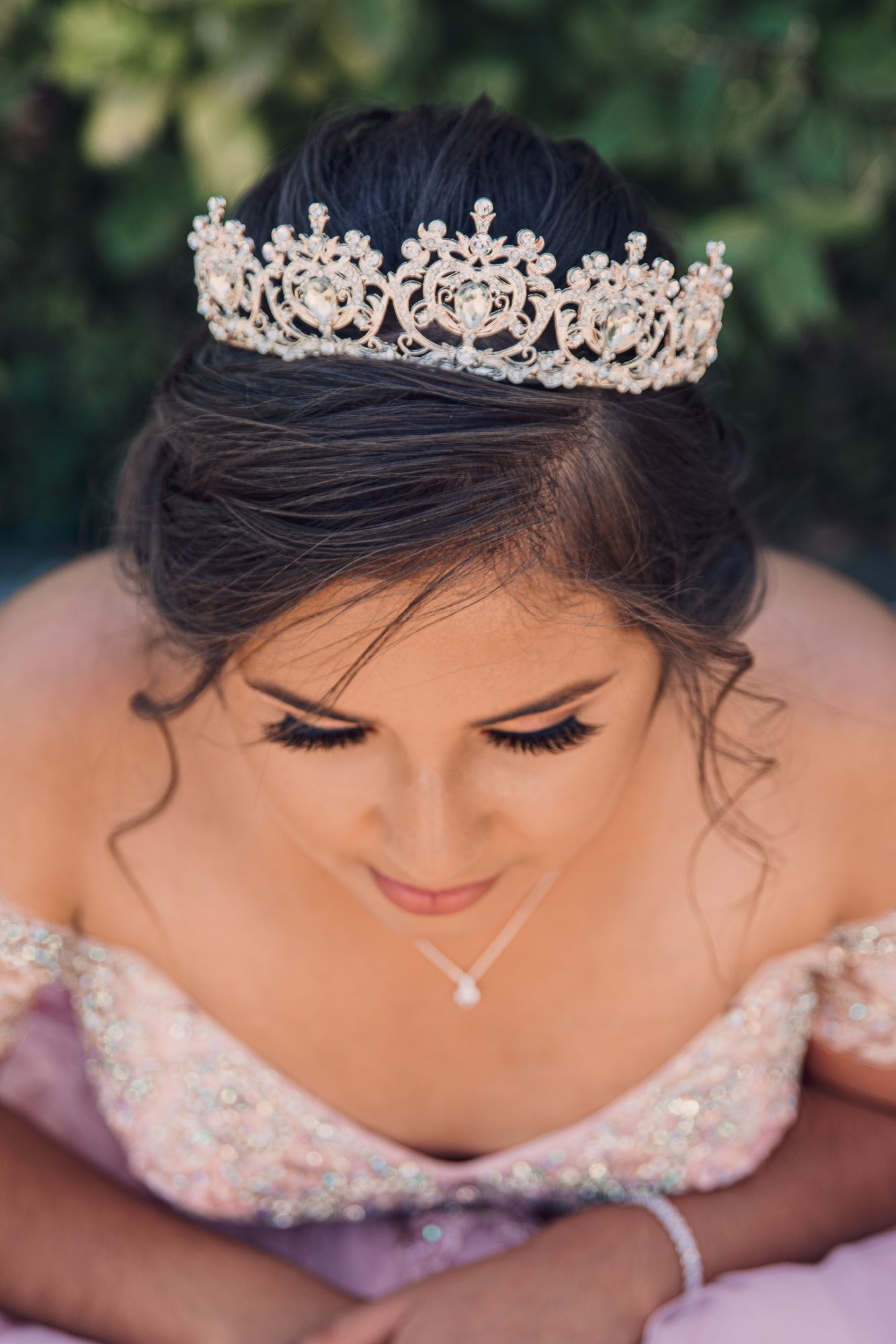 A delicate crystal tiara adorns a bridal hairstyle with soft curls against a natural outdoor backdrop.