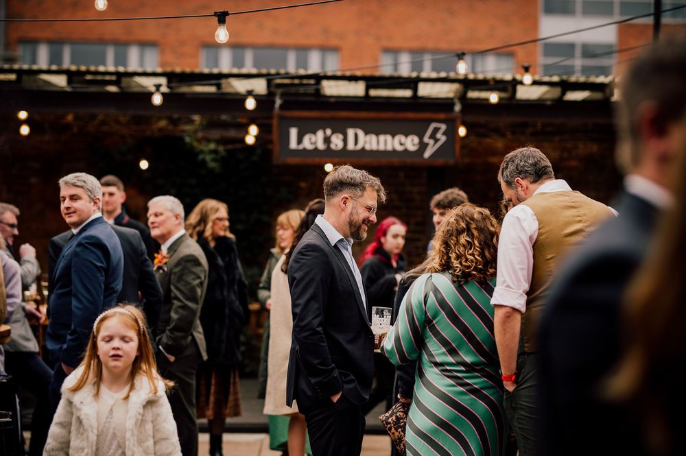 Wedding guests mingle and socialize at an evening reception under warm lighting and a 'Let's Dance' sign.