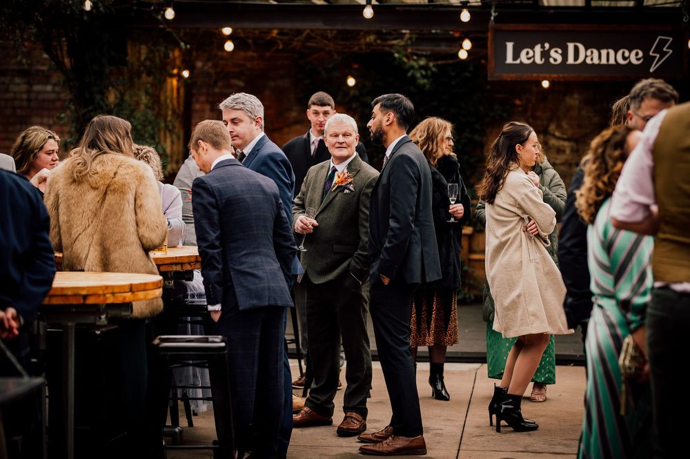 Guests gather at a rustic indoor wedding venue with string lights and a 'Let's Dance' sign overhead.