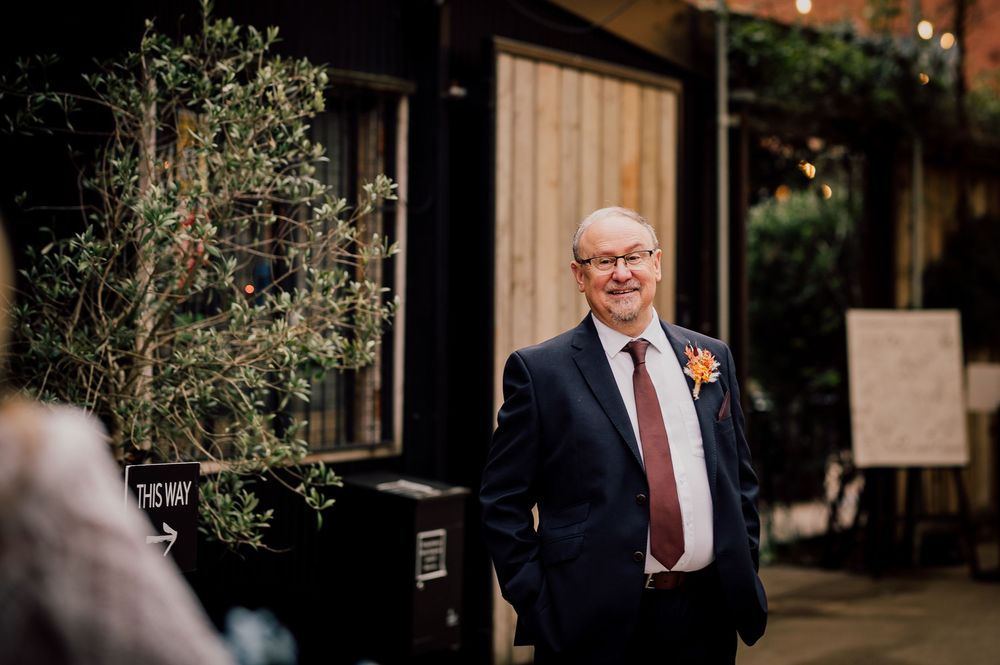A person in a navy suit and red tie smiles warmly while standing outside a building with greenery in the background.