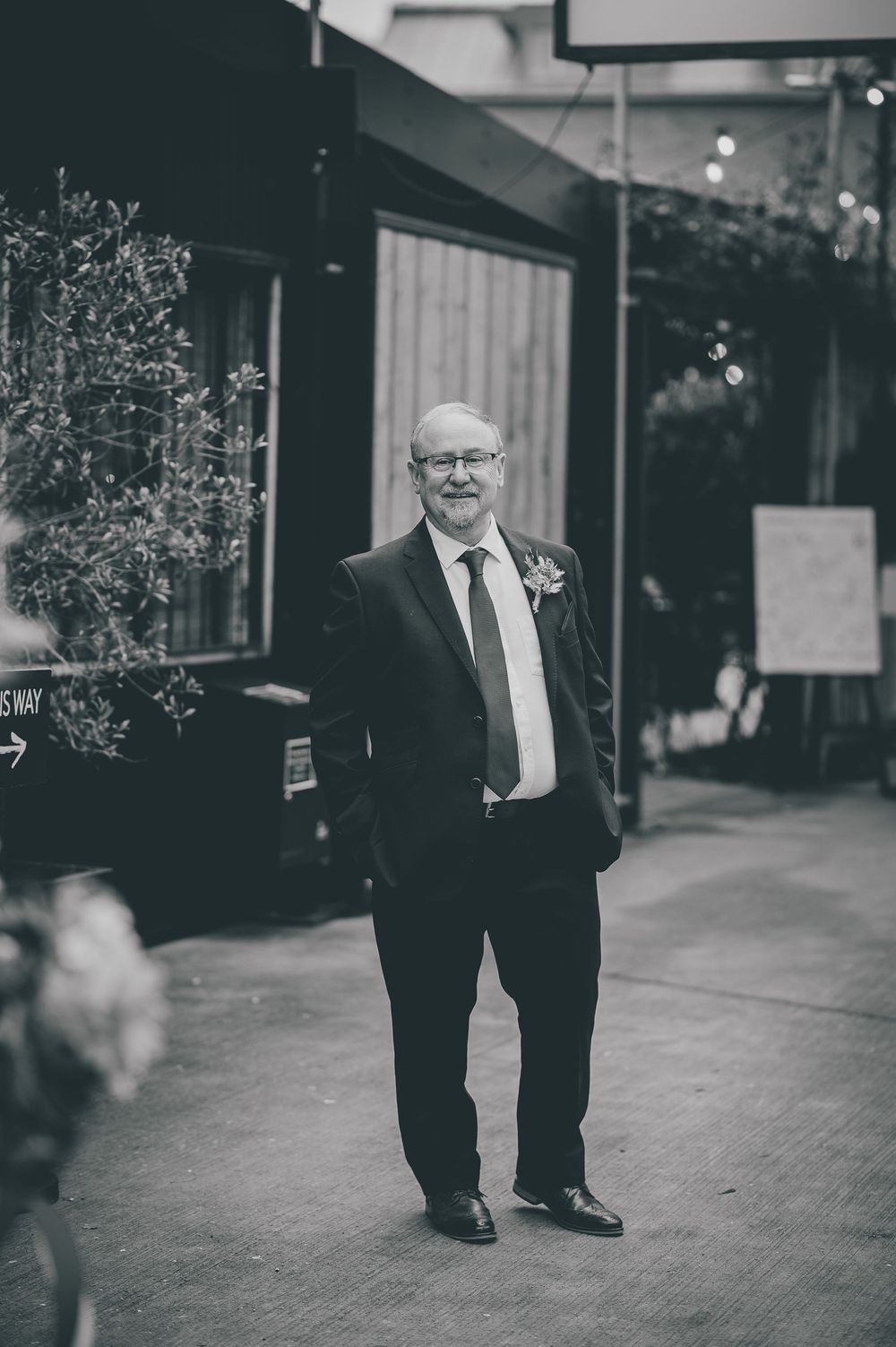 A person wearing a dark suit and boutonniere smiles while standing on a sidewalk outside a building.