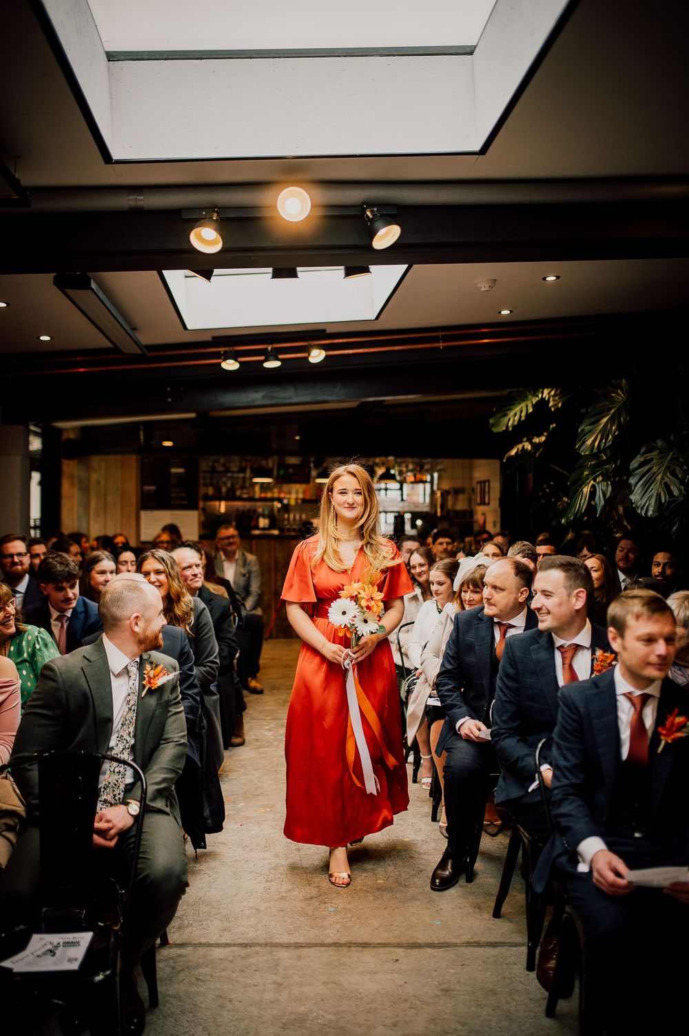 A bridesmaid in a vibrant red dress walks down the aisle during an indoor wedding ceremony while guests look on.